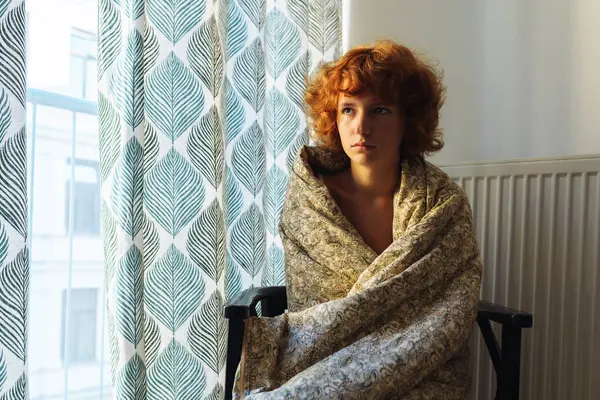 young woman, sleepy, red-haired, curly, wrapped in blanket, sitting on chair, near heating radiator, frozen