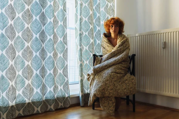 young woman, sleepy, red-haired, curly, wrapped in blanket, sitting on chair, near heating radiator, frozen