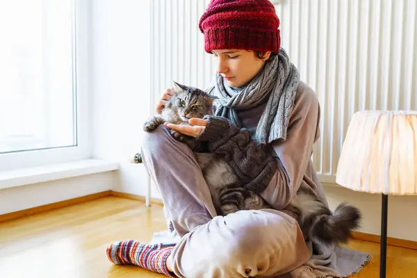 young girl in warm clothes, knitted warm socks, wearing hat and mittens, sitting on floor near radiator, hugging cat, warming himself, with floor lamp turned on nearby