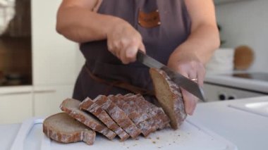 Kahverengi önlüklü tanınmayan bir kadın tahtanın üzerinde çavdar ekmeği dilimliyor.