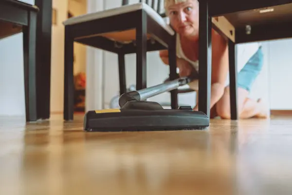Woman cleaning apartment with parquet floor with vacuum cleaner. woman crouching using vacuum cleaner under dining table at home. vacuum cleaner under