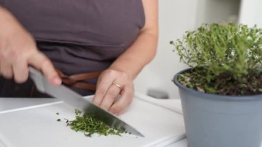 Kahverengi önlüklü kadın hemen ve profesyonel olarak mutfak bıçağıyla tahta keserek bitki doğruyor. Baharat hazırlama, kekik baharatları
