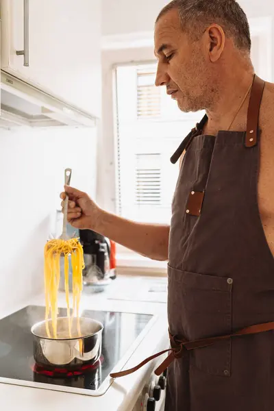 man cooks pasta spaghetti at home in kitchen. man cook in brown apron, tries readiness spaghetti, holds spaghetti on special spoon over boiling water. Italian home cooking concept