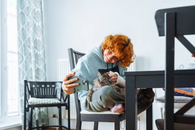 Genç kız, kızıl saçlı, çekici, kolu kırık, kolu alçıda, akıllı telefon kullanarak iletişim kuruyor, video konferans yoluyla iletişim kuruyor, oturma odasında sandalyede oturuyor.