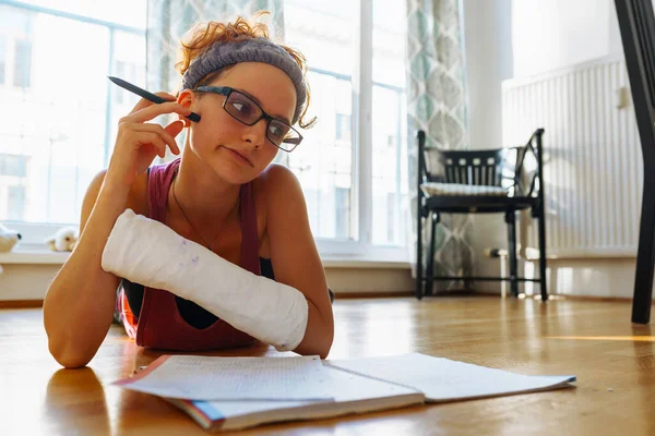 teenage girl with broken arm in cast, wearing glasses, lies on parquet floor, resting head on hand, studying, writing notes in notebook, doing homework, at home