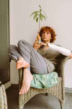 Genç kız, kareli yeşil gömlekli, kıvırcık kızıl saçlı, alçılı kolu kırık, balkondaki hasır bahçe sandalyesinde elinde çiçekle oturuyor. Elinde cam kavanozda filizlenmiş avokado.