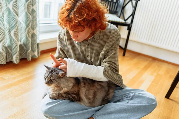 teenage girl, with broken arm in cast, in checkered shirt, sitting on parquet floor at home, holding domestic fluffy gray cat in arms. girl plays, teases pet, jokes