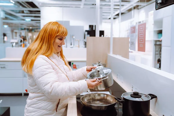 red-haired middle-aged woman in kitchen appliance store chooses kitchen utensils pot, frying pan.