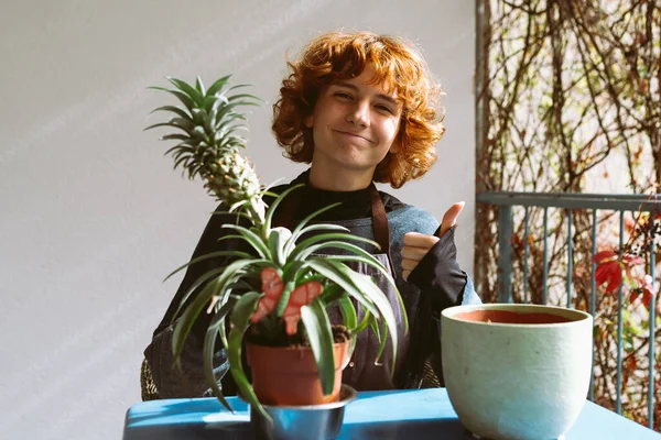 red-haired curly teenage girl, on home balcony or terrace, transplants tropical exotic plant, pineapple, into fertile soil. Home gardening, caring for house plants. replanting plant in large flowerpot