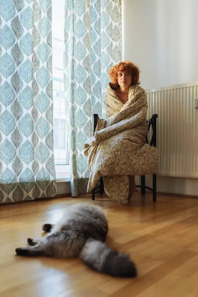 Girl hugging cat, sitting in blanket home, frozen. teen barefoot, red-haired, curly, morning, wrapped in blanket, sits hugging domestic cat, near heating radiator, large window covered with curtains.