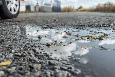 Taşra yolundaki su birikintisi, gökyüzünün yansıması, düşen sarı yapraklar ve araba tekerleği. Sonbahar depresif gerçekliği. Yol onarımı