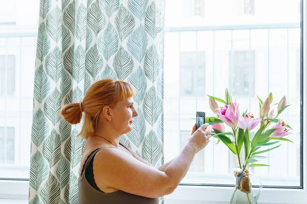 middle-aged woman with red hair tied in bun takes photo bouquet lilies standing in jug on windowsill at home using smartphone