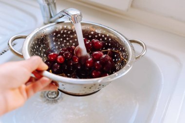 Kadınların elleri mutfak musluğundan akan suyun altında bir demet kırmızı üzümü kevgire batırır.