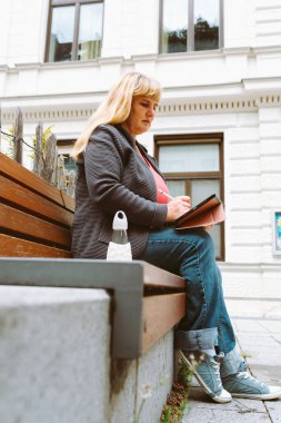 Orta yaşlı, uzun sarı saçlı, tabletli bankta oturan çekici bir kadın portresi.