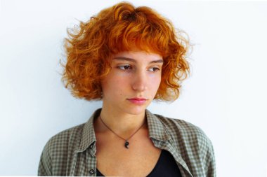 portre kızıl saçlı, kıvırcık saçlı, çekici, yüzünde duygular olan, erkek gömleği giymiş, arka plan duvarına yaslanmış genç kız.