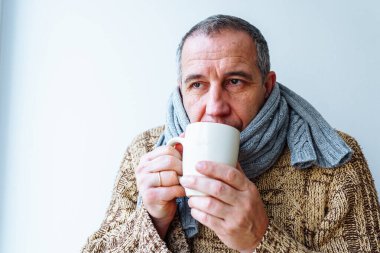Orta yaşlı bir adam portresi, sıcak örgü eşarp, süveter, sıcak içecek, evdeki ısıtma kaloriferinin yanında oturan. Soğuk, sağlık durumu, soğuk ev kavramı