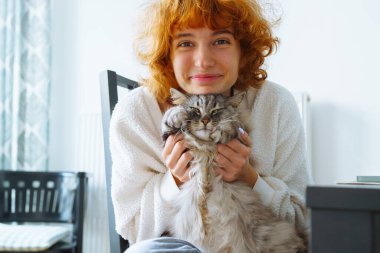 Portre kızıl saçlı, çekici, beyaz kazaklı, gülümseyen genç kız. Evdeki tüylü evcil kediye sarılıyor.