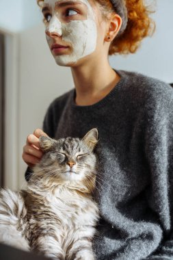 Gri tüylü tekir kedi kollarında oturuyor. Kızıl saçlı, saç bandıyla bağlanmış, kozmetik kilden maske takıyor.