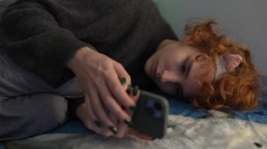 Saçında saç bandı olan genç bir kız, akşamları yatakta uzanıyor, cep telefonu kullanıyor, sosyal ağlara takılıyor.
