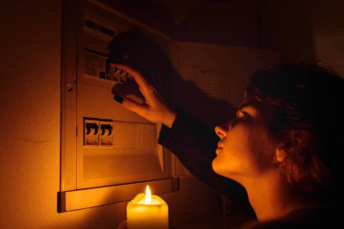 Tamamen karanlıkta mumları olan genç bir kadın elektrik kesintisi sırasında sigorta kutusunu veya elektrik dağıtım panelini inceliyor. Elektrik yok, elektrik yok.