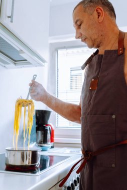 Adam evde, mutfakta makarna pişiriyor. Kahverengi önlüklü bir adam spagettiyi dener, spagettiyi kaynamış suyun üzerinde özel kaşıkta tutar. İtalyan ev yemeği konsepti