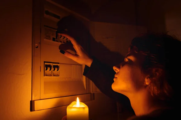 Tamamen karanlıkta mumları olan genç bir kadın elektrik kesintisi sırasında sigorta kutusunu veya elektrik dağıtım panelini inceliyor. Elektrik yok, elektrik yok.