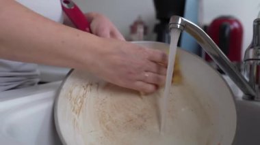 Kırmızı manikürlü eller kirli, kızarmış tava ve mutfak lavabosunda yağlı kahverengi lekeler. Bulaşık deterjanıyla tavayı yıkamak için bez kullanan kadın.