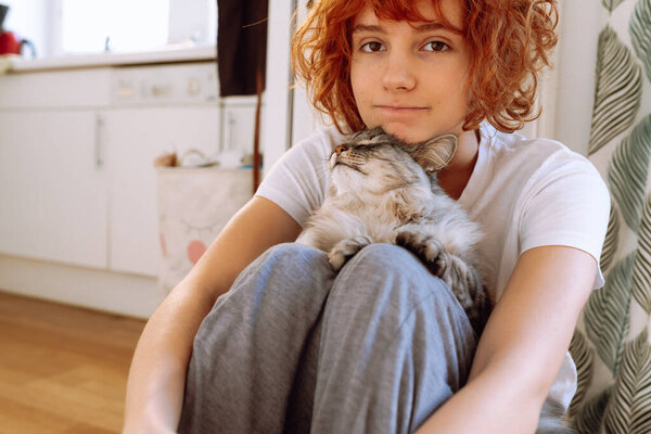 funny red-haired teenage girl sitting on floor near heating radiator at home hugs and kisses a purring Maine Coon cat. Friendship between pet and its owner
