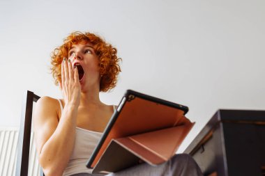 Tişörtlü, pijama pantolonlu, tablet kullanarak yemek masasındaki sandalyeye oturan kızıl saçlı genç kız. hafta sonu sabahı