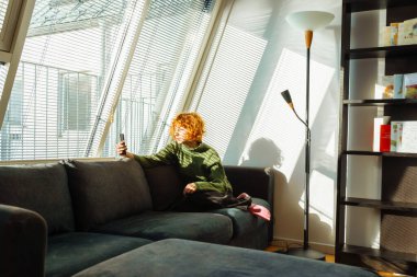 young attractive woman, student, sits on comfortable sofa, near large attic window, uses telephone, in bright sunny room, with parquet flooring. clipart