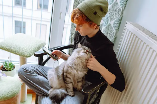 teenage girl sits on chair by window, wearing hat, holding domestic cat with mustache, using phone comfortably.