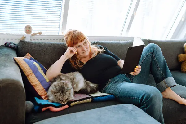 middle-aged woman with red hair sits on sofa, near large window, reading book, with gray fluffy cat lying next to her. side view home life