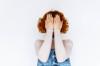 Çekici, kıvırcık saçlı, kot tulumlu, bej diz boyu çoraplı, yüzünde, beyaz arka planda duyguları olan genç, çekici bir kız portresi. Yüzü ellerle kaplı