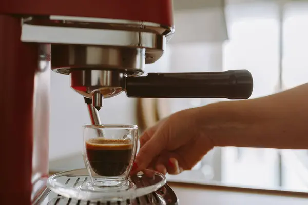 womans hand holding glass cup with fresh aromatic coffee. Espresso coffee poured into glass transparent cup from coffee machine, against blurred background morning light, in home kitchen