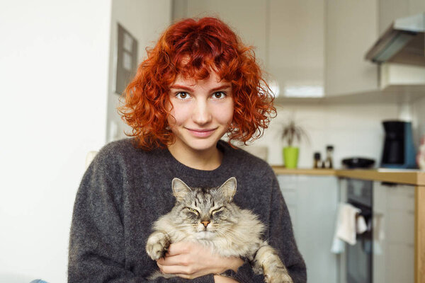 funny red-haired teenage girl sitting at home against blurred kitchen background hugs and kisses purring Maine Coon cat. Friendship between pet and its owner