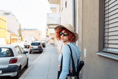 Portre genç bir kadın, çekici bir genç kız, kızıl kıvırcık saçlı, yaz şapkası ve güneş gözlüğü takan, Avrupa sokaklarında yürüyen, turizm konsepti, öğrencilik,