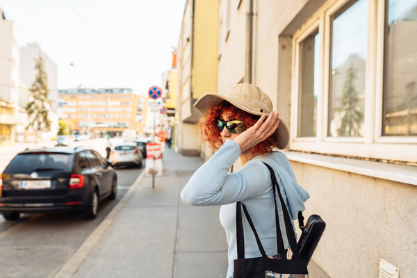 portrait young woman, an attractive teenage girl, student, with red curly hair, wearing summer hat and sunglasses, walking along European streets, tourism concept, studenthood,
