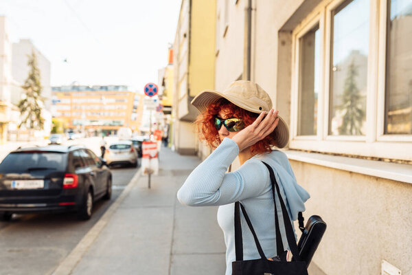 portrait young woman, an attractive teenage girl, student, with red curly hair, wearing summer hat and sunglasses, walking along European streets, tourism concept, studenthood,