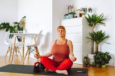 Orta yaşlı bir kadın, sarışın, kıvrımlı, ev bitkileriyle dolu parlak geniş bir odada yoga yapıyor, ev içi, vücut pozitifliği