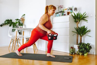 Orta yaşlı bir kadın, sarışın, kıvrımlı, ev bitkileriyle dolu parlak geniş bir odada yoga yapıyor, ev içi, vücut pozitifliği