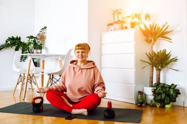 Orta yaşlı bir kadın, sarışın, kıvrımlı, ev bitkileriyle dolu parlak geniş bir odada yoga yapıyor, ev içi, vücut pozitifliği