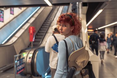 Portre çekici kızıl saçlı genç kadın, pantolonu, mavi kazağı, şapkası, metroda veya istasyonda, yürüyen merdivenin yanında.