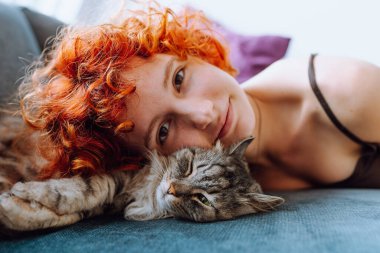 portre kızıl saçlı genç kız gri tüylü kedi, pozitif ruh halinde, rahat bir kanepede uzanıyor.