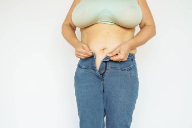 Kot pantolonlu ve üstte orta yaşlı bir kadın göbeğini, sarkık tenini ve kilosunu gösteriyor. Vücut pozitifliği, vücut yaşlanmaları, vücut kusurları, doğum lekeleri ve kıllar.