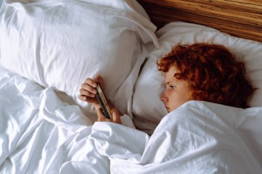 Çekici kızıl saçlı, kıvırcık saçlı, beyaz bir yatakta uzanmış, akıllı telefon kullanıyor.