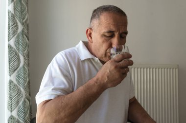 Beyaz polo giyen orta yaşlı bir adam, bardaktan su içiyor, yakın plan. Soluk tonlama, sağlık konsepti, insan hastalığı.