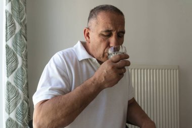 Beyaz polo giyen orta yaşlı bir adam, bardaktan su içiyor, yakın plan. Soluk tonlama, sağlık konsepti, insan hastalığı.