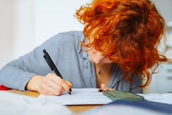 stock image young woman, student, with red curly hair, fills out documents sitting at table at home, in foreground is passport citizen of Ukraine. citizenship concept, temporary asylum in European countries