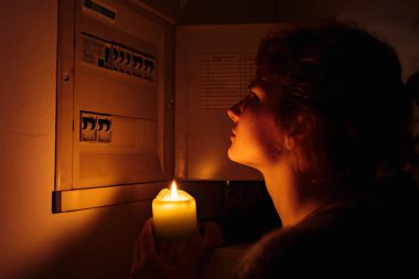 Tamamen karanlıkta mumları olan genç bir kadın elektrik kesintisi sırasında sigorta kutusunu veya elektrik dağıtım panelini inceliyor. Elektrik yok, elektrik yok.