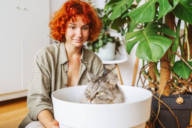 Evcil hayvanınla bahçeyle uğraşıyorsun. Yerel gri tüylü kedi saksının içinde oturuyor, genç kızıl saçlı kadın yanında oturuyor, komik bir durum. Saksı bitkilerini taze toprağa ekiyorum.
